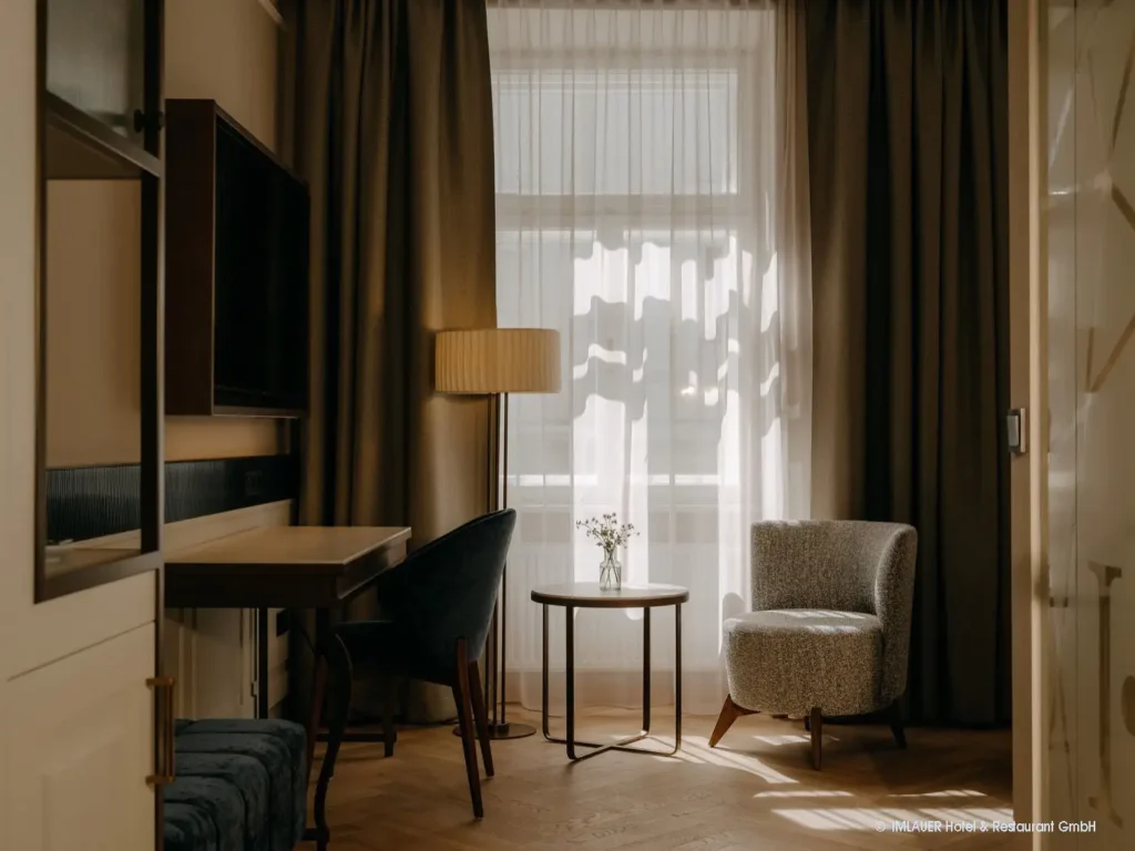 01_ImlauerPalaisMirabell_Copyright IMLAUER Gemütliche Sitzecke mit Sessel, Tisch und Stehlampe vor einem großen Fenster mit Vorhängen im Imlauer Palais Mirabell