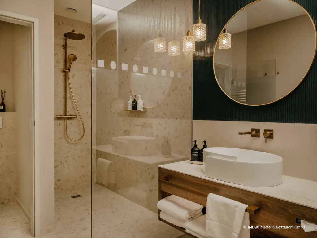 03_ImlauerPalaisMirabell_Copyright IMLAUER Modernes Badezimmer mit begehbarer Dusche, rundem Spiegel, Holzwaschtisch und goldfarbenen Armaturen im Imlauer Palais