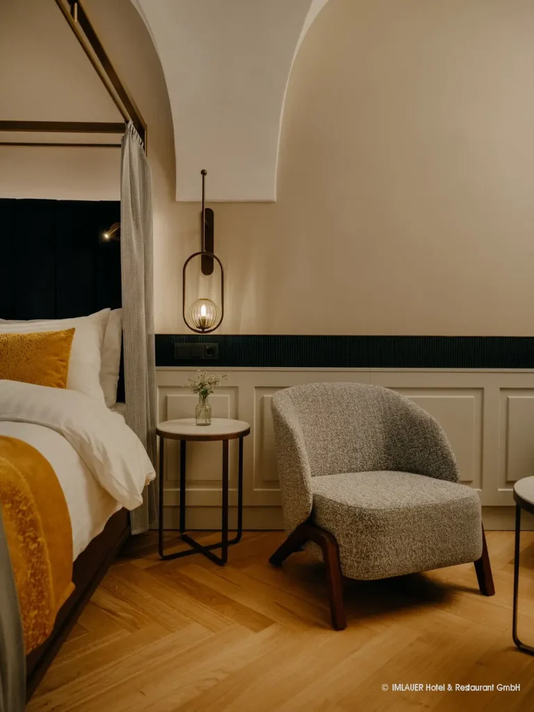 06_ImlauerPalaisMirabell_Copyright IMLAUER Gemütliche Hotellounge mit grauem Sessel, kleinem Beistelltisch, Bett mit gelben Kissen und moderner Wandlampe im Imlauer