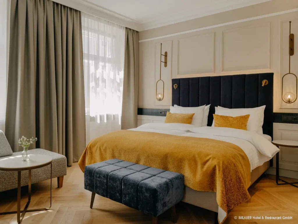 07_ImlauerPalaisMirabell_Copyright IMLAUER Modern eingerichtetes Hotelzimmer mit Doppelbett, gelber Bettdecke, dunkelblauem Polsterhocker und Sitzbereich am Fenster