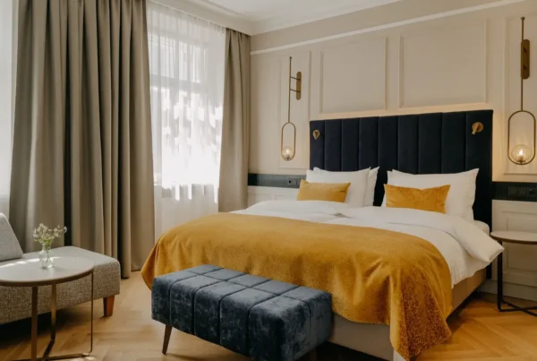 Modern eingerichtetes Hotelzimmer mit Doppelbett, gelber Bettdecke, dunkelblauem Polsterhocker und Sitzbereich am Fenster